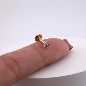 May include: A gold-colored lip piercing stud with a flat disc backing and a straight post. The stud features three small, green gemstones arranged in a triangular shape. The jewelry is shown on a finger.