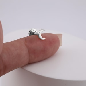 Może przedstawiać: Srebrny kolczyk labret z motywem półksiężyca. Kolczyk ma płaską, okrągłą podstawę z wygrawerowanymi oznaczeniami. Księżyc jest gładki i zakrzywiony, tworząc minimalistyczną i niebiańską estetykę.