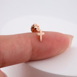 May include: A gold-toned cross stud earring with a small, round backing. The earring is shown on a finger.