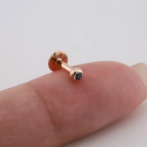 May include: A small, gold-colored stud earring with a dark blue gemstone. The earring has a flat, circular backing and a post. The gemstone is set in a rounded bezel.