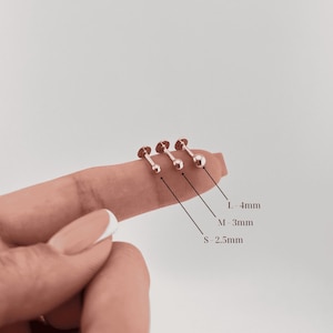 Puede incluir: Se muestran tres pendientes de botón de color oro rosa con respaldos de disco plano y bola. Los tamaños están etiquetados: S - 2,5 mm, M - 3 mm y L - 4 mm. Los pendientes se muestran en un dedo.