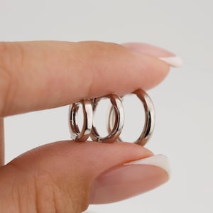 Puede incluir: Tres pendientes de aro plateados de diferentes tamaños se sostienen entre los dedos. Los pendientes tienen una superficie pulida y reflectante, y un diseño sencillo. El pendiente más pequeño es de estilo huggie, mientras que el más grande es un aro de tamaño mediano.
