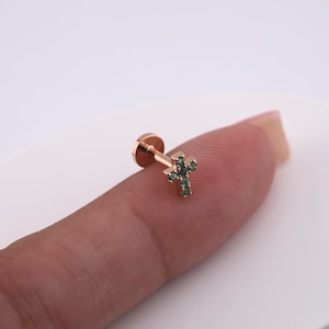 May include: A small, gold-colored stud earring featuring a cross design. The cross is adorned with small, green gemstones. The earring is shown on a finger against a white background.