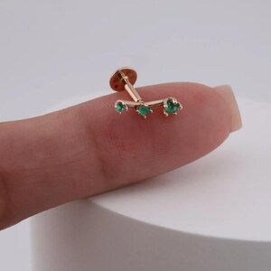 May include: A gold-colored ear stud with three green gemstones. The stud has a flat, round base and a curved bar with three emerald-colored stones. The gemstones are set in a flower-like shape.