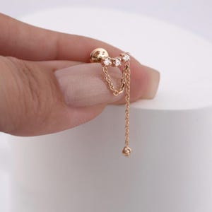 Puede incluir: Un pendiente dorado con tres pequeñas piedras preciosas transparentes en fila. Dos delicadas cadenas cuelgan de las piedras preciosas, con un pequeño dije cuadrado al final de la cadena más larga. El fondo es blanco.