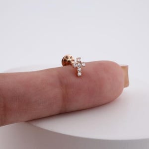 May include: A small, rose gold-colored earring with a cross design. The cross is adorned with several small, clear gemstones. The earring post is visible, and the background is a soft, neutral color.