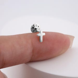 May include: A small, silver-colored cross earring. The earring features a simple cross design and a flat backing. The letters "L" and "R" are visible on the backing. The earring is shown on a finger.