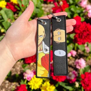 May include: Two black keychains with a white background. One keychain features a cartoon character with yellow hair and a red and yellow suit. The other keychain features the same character's face with the word "OK" in a speech bubble.