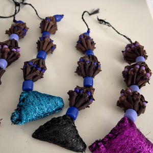 May include: Close-up of several decorative beaded tassels. Each tassel features a cluster of brown cloves, blue beads, and a colorful fabric accent in teal, black, and purple. Black string is attached to the top of each tassel.