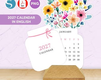 May include: A 2027 calendar in English featuring a jar-shaped cover with a pink bow and a monthly page for January. The calendar is decorated with a watercolor floral arrangement in yellow, pink, and blue tones, and it rests on a wooden base.