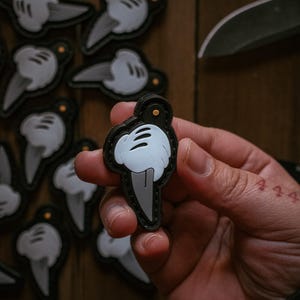 May include: A black and white patch featuring a cartoon hand holding a knife. The patch has a black border and a small gold accent. Several similar patches are visible in the background.