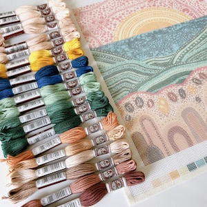 May include: A close-up of a needlepoint canvas featuring a colourful landscape design. The canvas is surrounded by a selection of DMC embroidery floss spools in various colours, including green, blue, yellow, orange and brown.