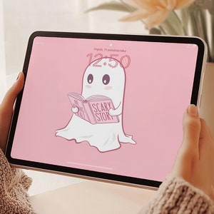 Puede incluir: Una tableta rosa con un fantasma leyendo un libro que dice "Scary Story" en la portada. El fantasma es blanco y tiene una cara rosa con ojos negros. La tableta está sobre una mesa de madera con un ramo de flores en el fondo.