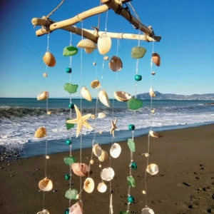 Puede incluir: Móvil de temática costera con conchas marinas, vidrio de mar y estrellas de mar suspendidas de una rama de madera natural. Las conchas varían en tamaño y forma, con tonos beige, blanco y marrón. Cuentas turquesas y hilo transparente completan el tema playero.