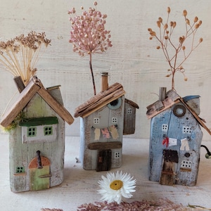 Peut inclure: Trois maisons miniatures en bois flotté, chacune peinte dans des couleurs différentes et décorées de détails comme des fenêtres, des portes et une cheminée. Les maisons sont disposées sur une surface blanche avec des fleurs séchées et une seule marguerite blanche.