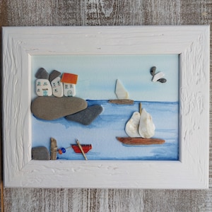 May include: A framed watercolor painting depicting a seascape with a small village of houses on a rocky shore, a sailboat on the water, and a small red boat docked at a pier. The painting is made with sea glass and other natural materials.
