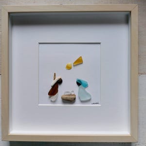 Può includere: Un'opera d'arte incorniciata che rappresenta la scena della Natività utilizzando vetro di mare e legno di deriva. L'opera presenta una figura marrone e bianca di Maria, una figura blu di Giuseppe e una piccola figura bianca di Gesù bambino in una mangiatoia fatta di legno di deriva. L'opera d'arte è su uno sfondo bianco con una stella gialla sopra le figure.