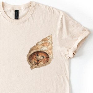 May include: A light beige t-shirt featuring a detailed illustration of a hermit crab inside a seashell. The artwork is in shades of brown and beige, with the crab's eyes visible. The shirt has a classic crew neck and short sleeves.