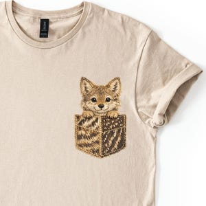 May include: A beige short-sleeved t-shirt featuring a graphic of a small fox peeking out from a faux pocket. The pocket design has a brown and tan patterned texture, and the fox has detailed fur, large eyes, and pointed ears.