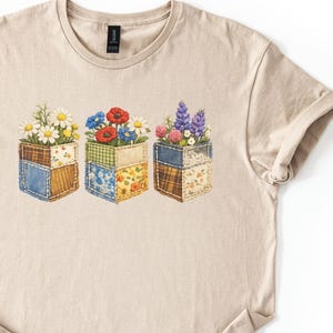 May include: A beige t-shirt with a floral design. The graphic depicts three square flower boxes, each filled with different flowers like daisies, poppies, and lavender. The boxes have a patchwork design in blue, green, and yellow.