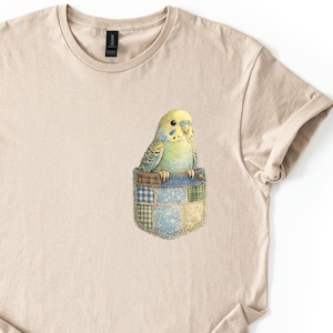 May include: Beige t-shirt featuring a colorful illustration of a parakeet in a patchwork pocket. The parakeet has yellow and green feathers. The pocket is made of blue, green, and yellow fabric squares. The shirt has short sleeves and a crew neck.