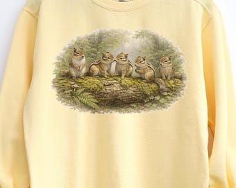 Könnte beinhalten: Hellgelbes Sweatshirt mit einer detaillierten Illustration von fünf Streifenhörnchen auf einem moosbewachsenen Baumstamm in einem Wald. Die Streifenhörnchen sind in verschiedenen Posen dargestellt.