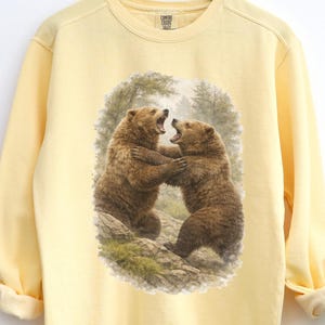 May include: A light yellow sweatshirt featuring a detailed illustration of two brown bears in a forest setting. The bears are depicted in a dynamic pose, seemingly interacting with each other. The sweatshirt has long sleeves and a crew neck.