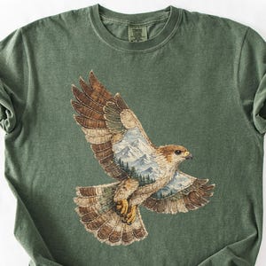 May include: Green t-shirt with a detailed illustration of a hawk in flight. The hawk's wings are spread, revealing a mountain and forest scene within. The shirt is a solid green colour.
