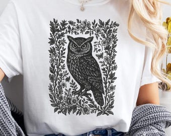 Könnte beinhalten: Weißes T-Shirt mit einer schwarz-weißen Illustration einer Eule, umrahmt von Blumen und Blättern. Das Design ist mittig platziert. Das T-Shirt wird mit einer blauen Jeans kombiniert.
