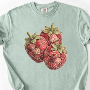 Op de afbeelding: Lichtgroen T-shirt met een afbeelding van drie rode frambozen. De frambozen hebben groene stelen en bladeren, en een patchwork ontwerp met rode en witte stippen en bloempatronen.
