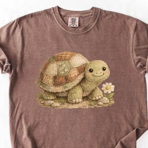 May include: A brown t-shirt with a cartoon turtle design. The turtle has a patchwork shell, a smiling face, and a daisy. The t-shirt has a crew neck and short sleeves. The t-shirt is a brown color.