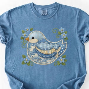 May include: A light blue t-shirt featuring an embroidered bird design. The bird is composed of various blue patterned fabrics and surrounded by blue flowers and green leaves. The shirt has rolled-up sleeves.