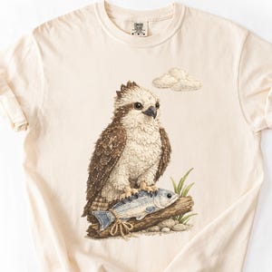 Op de afbeelding: Crèmekleurig T-shirt met een gedetailleerde illustratie van een roofvogel die op een boomstam met een vis zit. De vogel heeft bruine en witte veren en de vis is blauw en zilver. Een kleine wolk staat op de achtergrond.