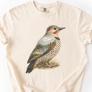 May include: Cream-colored t-shirt featuring a detailed illustration of a bird with a landscape design. The bird has a red head, black markings, and a speckled chest. The landscape design includes trees and mountains. The shirt is made of soft material.