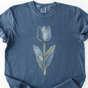 May include: A blue t-shirt with a denim tulip design. The tulip is made from denim patches with white stitching. The shirt has rolled-up sleeves. The t-shirt is a solid blue colour.