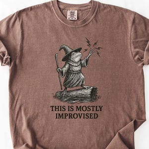 May include: A brown t-shirt featuring a whimsical illustration of a frog dressed as a wizard, holding a staff and casting a spell. The text "THIS IS MOSTLY IMPROVISED" is printed below the illustration. The shirt has a crew neck and short sleeves.