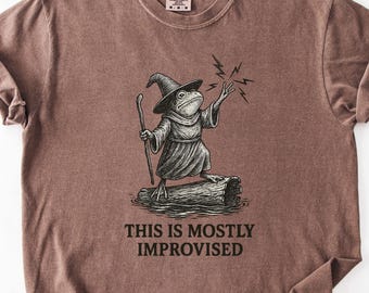 Op de afbeelding: Bruin t-shirt met een humoristische illustratie van een kikker-tovenaar, die een staf vasthoudt en een spreuk uitspreekt. De tekst "THIS IS MOSTLY IMPROVISED" staat eronder. Het shirt heeft een ronde hals en korte mouwen.