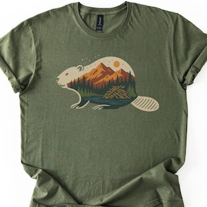 Könnte beinhalten: Olivgrünes T-Shirt mit einem Biber-Design. Die Biber-Silhouette enthält eine Landschaft mit Bergen, Bäumen, einem See und einer Sonne. Das Design ist in Braun-, Orange-, Grün- und Blautönen gehalten. Das T-Shirt hat hochgekrempelte Ärmel.