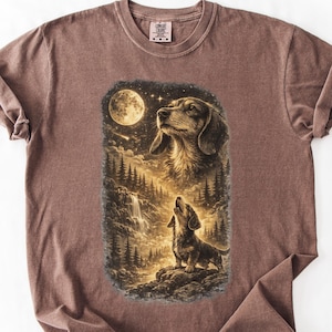 May include: A brown t-shirt featuring a detailed graphic of a dachshund howling at the moon in a forest scene. The design includes a waterfall, trees, and stars, with a second image of the dog's head. The shirt has a crew neck and short sleeves.