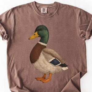 May include: A brown t-shirt featuring a detailed illustration of a mallard duck. The duck has a green head, brown chest, and a patchwork-style body with blue accents. The shirt has a crew neck and short sleeves.