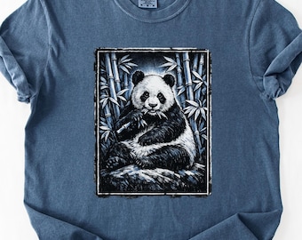 May include: A blue t-shirt featuring a black and white illustration of a panda bear eating bamboo. The panda is set against a backdrop of bamboo stalks. The shirt has short sleeves and a crew neck.