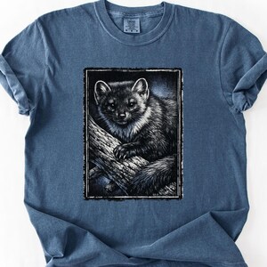 May include: A blue t-shirt featuring a black and white illustration of a small animal, possibly a marten or fisher, perched on a tree branch. The graphic is framed in a black rectangle.