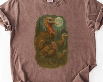 Puede incluir: Camiseta marrón con una ilustración detallada de pavos en un entorno forestal bajo la luna llena. El diseño incluye varios pavos en varias poses, con una paleta de colores marrón, verde y crema. La camiseta tiene cuello redondo clásico y mangas cortas.