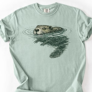 May include: A pale green t-shirt with a detailed illustration of a beaver swimming in water. The beaver has brown fur and is surrounded by concentric ripples. The shirt is made of a soft material.