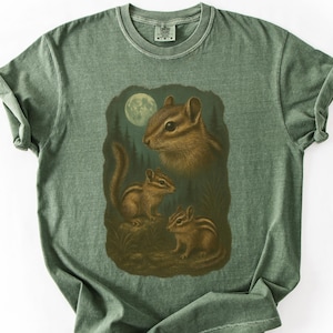 May include: Green t-shirt featuring a detailed illustration of three chipmunks under a full moon. The chipmunks are depicted in shades of brown and tan, with a forest backdrop. The shirt has a classic crew neck and short sleeves.