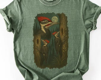 May include: Green t-shirt featuring a detailed illustration of a Pileated Woodpecker family. The design includes a large woodpecker with a red crest, two smaller woodpeckers, a full moon, and a forest backdrop. The shirt is made of soft material.