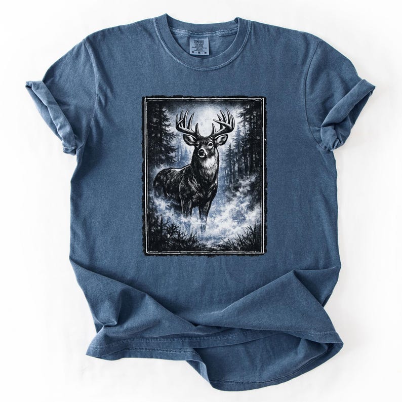 K&ouml;nnte beinhalten: Blaues T-Shirt mit einer Schwarz-Wei&szlig;-Illustration eines Hirsches in einem Wald. Der Hirsch hat ein gro&szlig;es Geweih und steht in einer nebligen Gegend. Das T-Shirt hat hochgekrempelte &Auml;rmel.