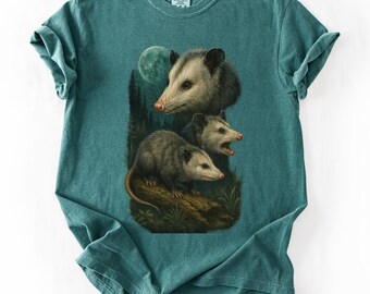 May include: Teal-colored t-shirt featuring a detailed illustration of three opossums. The opossums are depicted in a naturalistic style, with one looking forward, and two with open mouths. A full moon and forest scene are in the background.