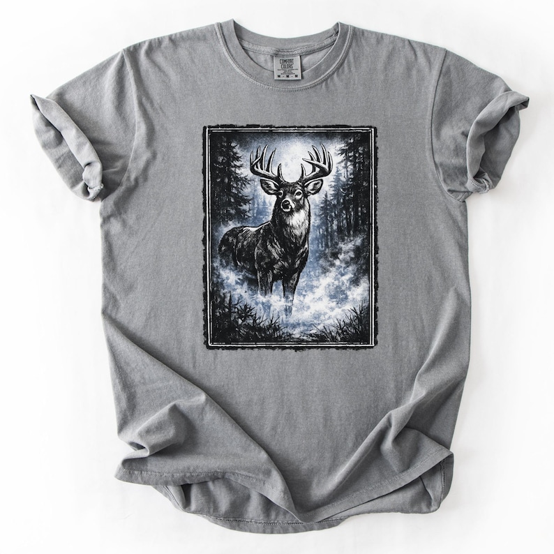 K&ouml;nnte beinhalten: Graues T-Shirt mit einer Schwarz-Wei&szlig;-Illustration eines Hirsches in einem Wald. Der Hirsch hat ein gro&szlig;es Geweih und steht in einer nebligen Gegend. Das T-Shirt hat einen Rundhalsausschnitt und kurze &Auml;rmel.
