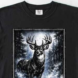 K&ouml;nnte beinhalten: Schwarzes T-Shirt mit einer detaillierten Illustration eines Hirsches mit gro&szlig;em Geweih. Die Grafik ist vor dem Hintergrund einer Waldszene platziert. Das T-Shirt hat einen Rundhalsausschnitt und das Comfort Colors-Logo ist sichtbar.
